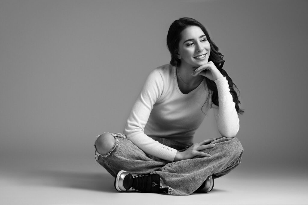 Jeune femme adolescence en séance photo, assise au sol, en jeans et pull, sourire aux lèvres, pour la création de son book