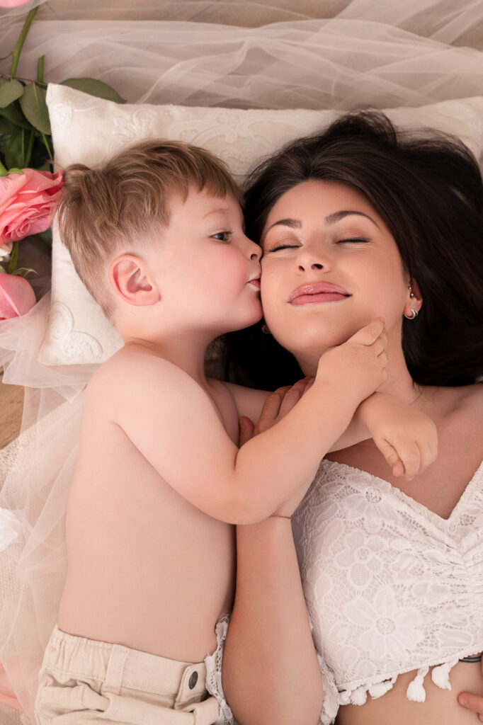 Petit garçon donnant un bisou sur la joue de sa maman allongée entourée de roses roses lors d'une séance photo Fête des Mères au Studio Evidence à Pusignan